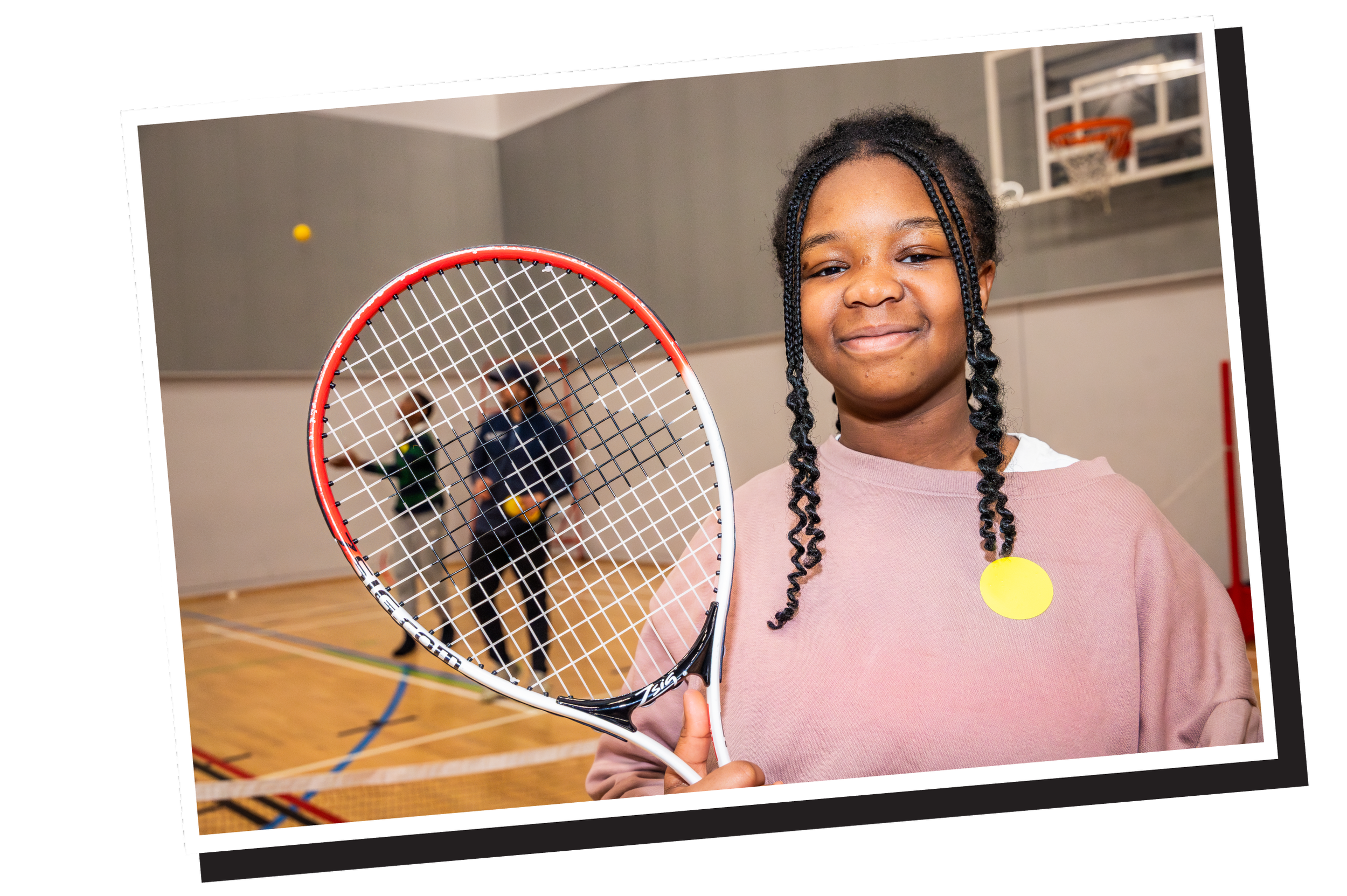 Young girl holding a tennis racket in an indoor gym with basketball hoop in background.