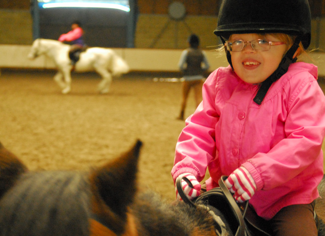 The image shows a child riding a horse with another rider in the background