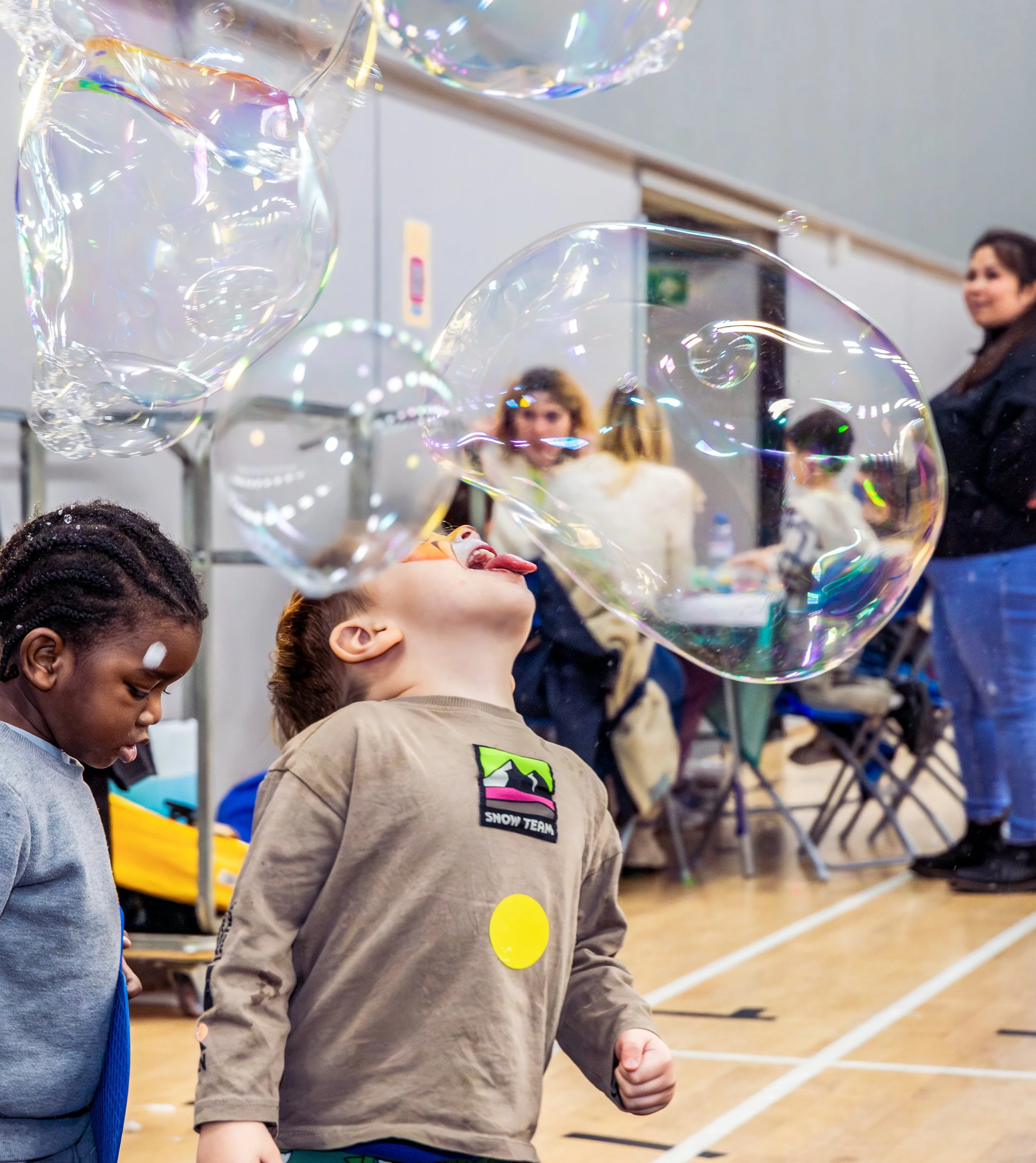 Two boys are playing with giant bubbles. One has his tongue stuck out and is trying to lick a bubble as it floats through the air.