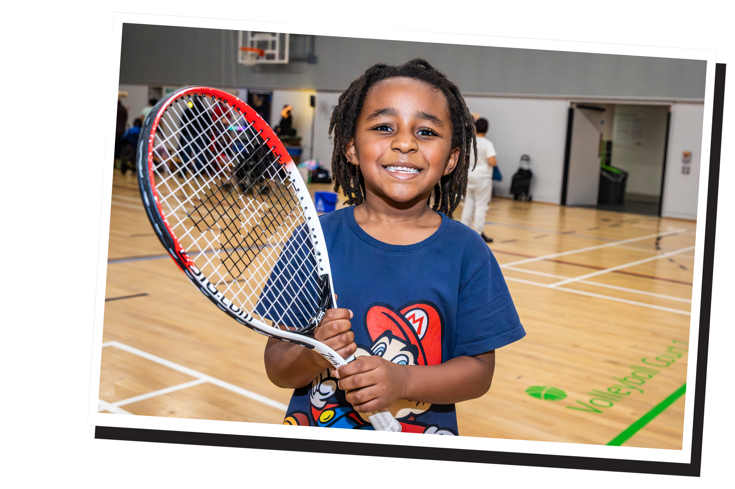 Young boy smiling and holding a tennis racket in an indoor gymnasium.