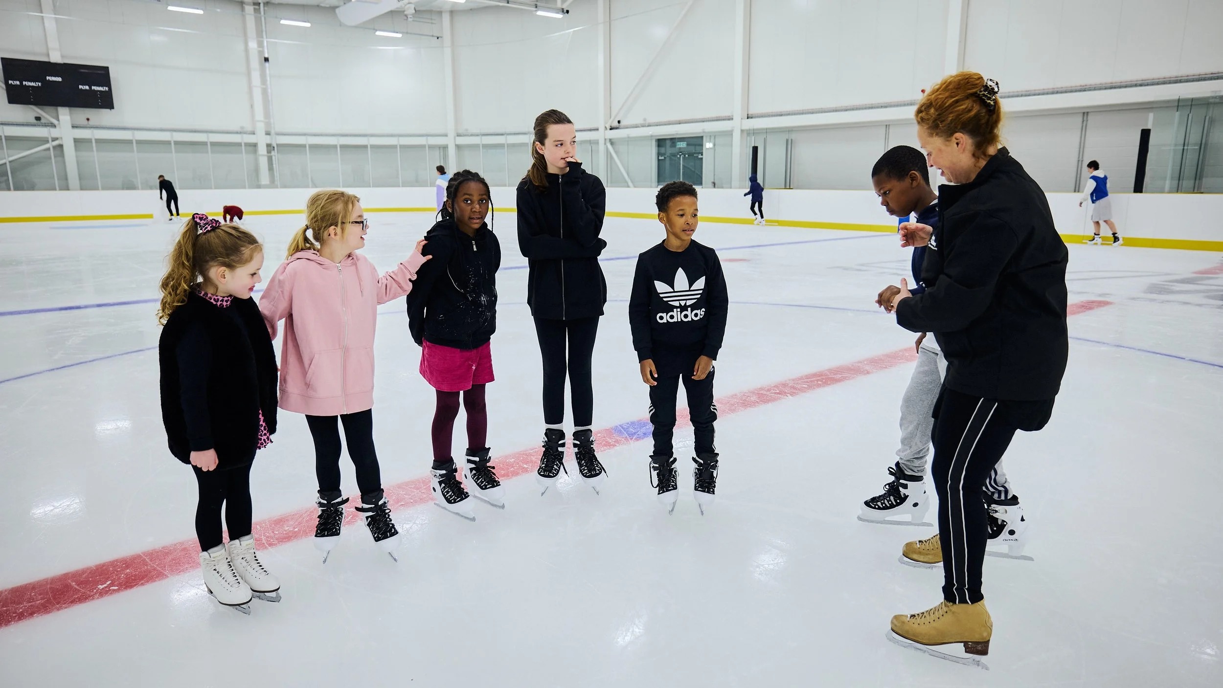 The image shows a group of children and young people standing on the ice with an instructor