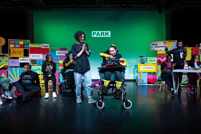 The image shows a group of young people on stage, some are seated, some are standing and their is a wheelchair user in centre stage