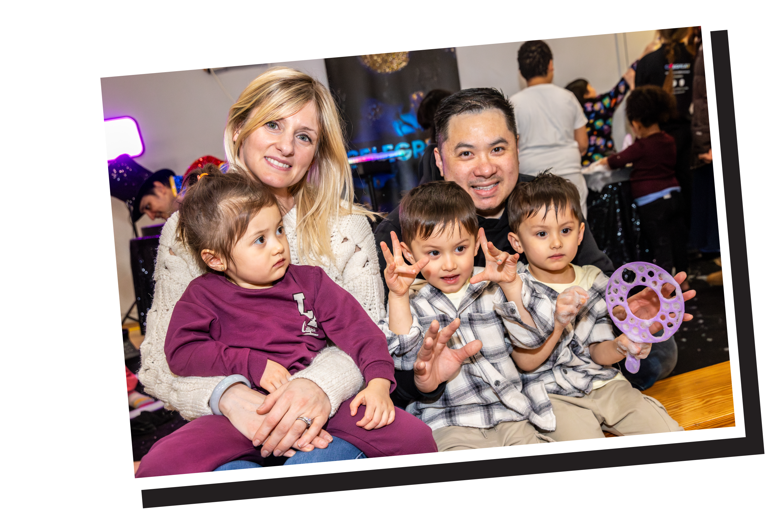 A group of people, including children and adults, sitting together at a gathering or event. The children are making hand gestures, and one is holding a purple toy or object. The background shows a dark wall with lights and other attendees.