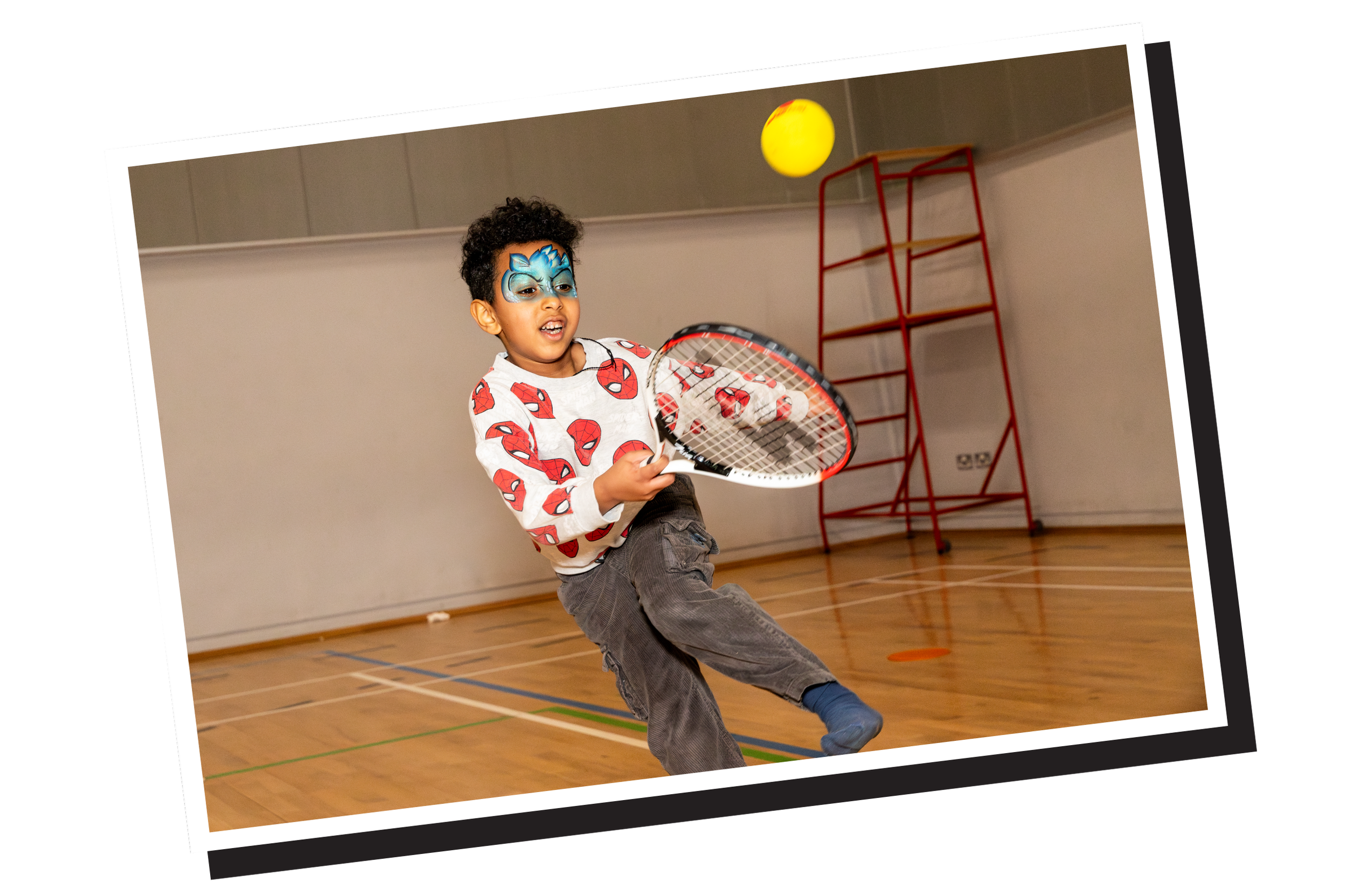 A young boy wearing face paint and a Spider-Man sweatshirt is playing tennis indoors, hitting a yellow tennis ball with a racket.