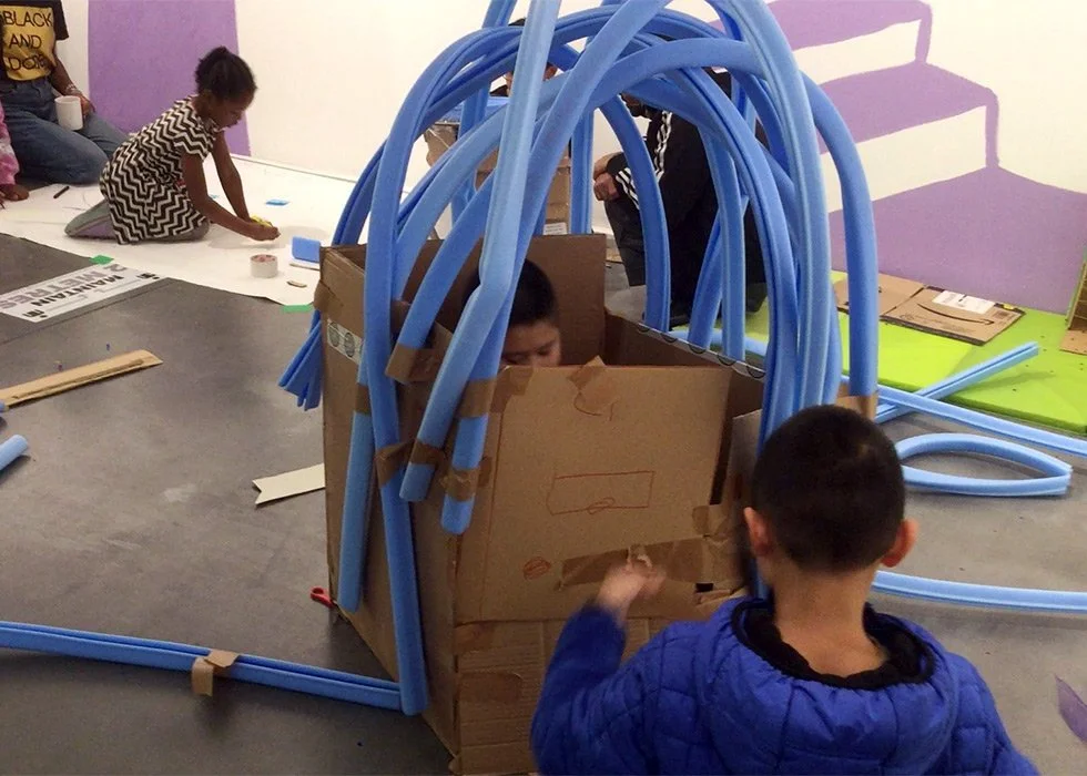 The image shows a group of children doing a creative project involving a large box which a child is sat inside and blue tubing above it