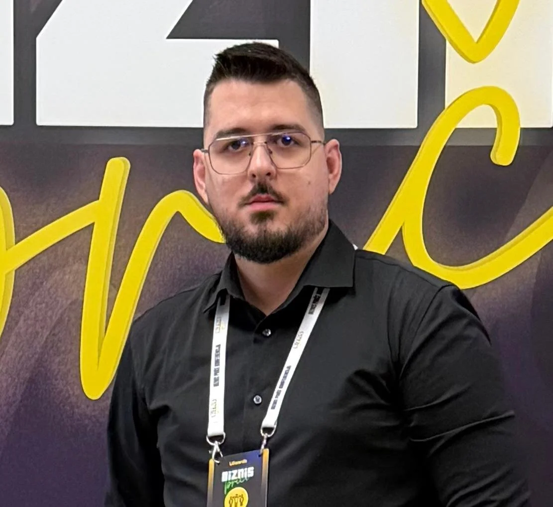 A man with glasses, a beard, and short hair posing inside an elevator with metallic walls, wearing a dark turtleneck and light gray blazer.