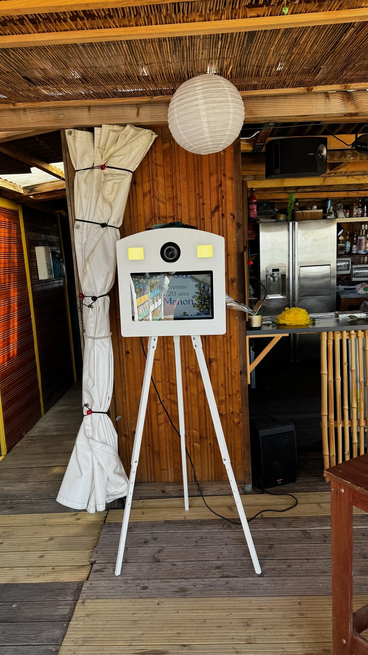 Photo of a white photo booth with a digital screen on a tripod stand in a wooden outdoor space. The screen displays a welcoming message in French for a 20th anniversary celebration. There is a paper lantern hanging from the ceiling, and a curtain tied to the side. In the background, there is a kitchen area with stainless steel appliances and various decorations.