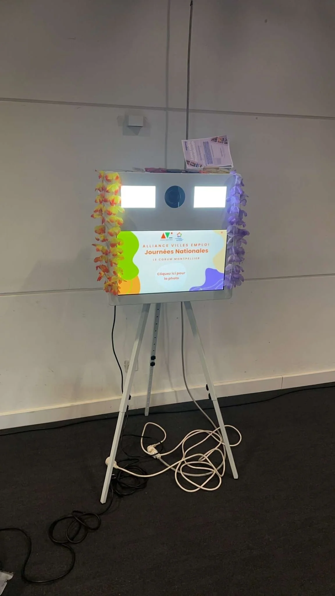 A photo kiosk with a colorful display screen that reads 'Alliance Villes Emploi! Journées Nationales' and has flower leis hanging from the sides. There are printed flyers on top and an extension cord with multiple plugs on the floor.