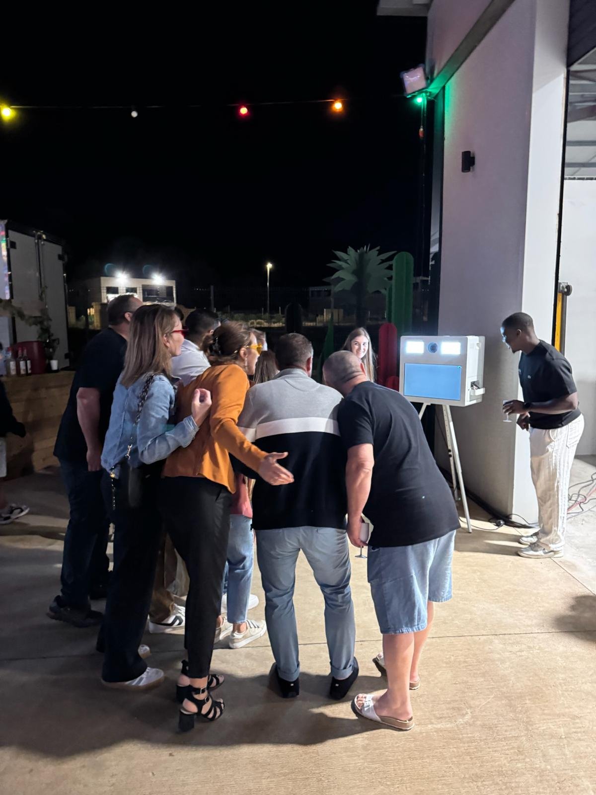 A group of people gathered around at an outdoor karaoke booth at night. They are watching a large screen displaying a woman singing. There is a man operating the karaoke machine on the right. The scene includes Christmas or festive lights and desert-themed decorations like cactus shapes.