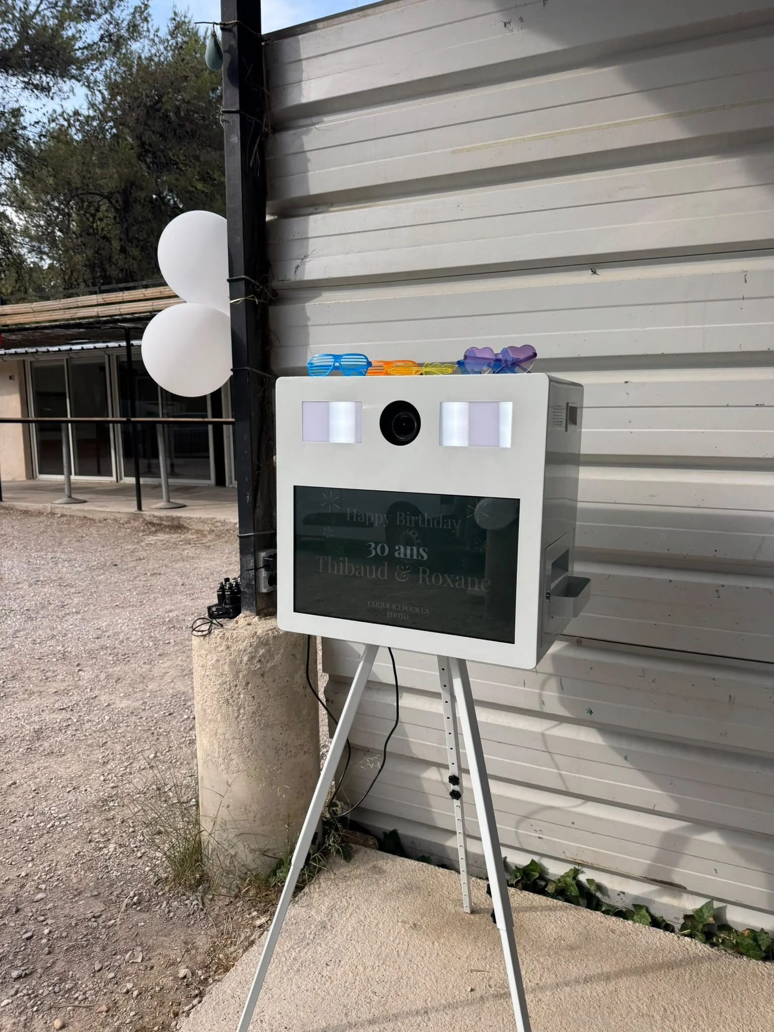 Outdoor photo of a photo booth with a sign reading "Happy Birthday 30 ans Thibaud & Roxane." The photo booth has sunglasses and heart-shaped glasses on top and is placed in front of a white metal building with trees in the background. White balloons are attached to a black pole nearby.