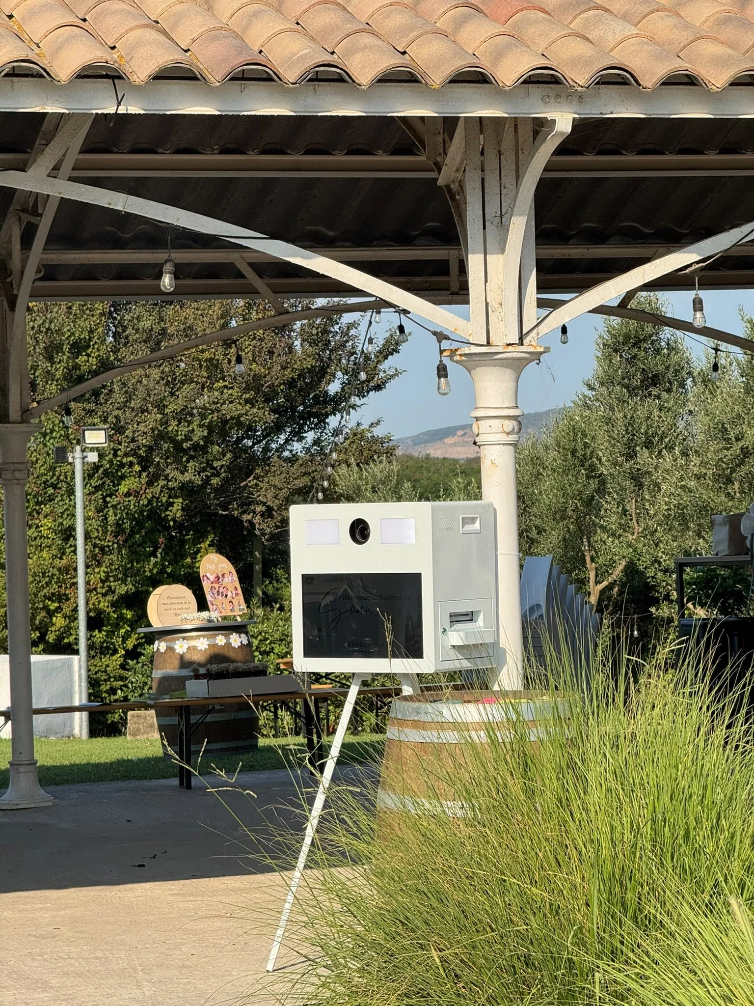 Outdoor scene showing a white vertical photo booth with a circular camera lens and screens, set under a patio with string lights, surrounded by trees and greenery.