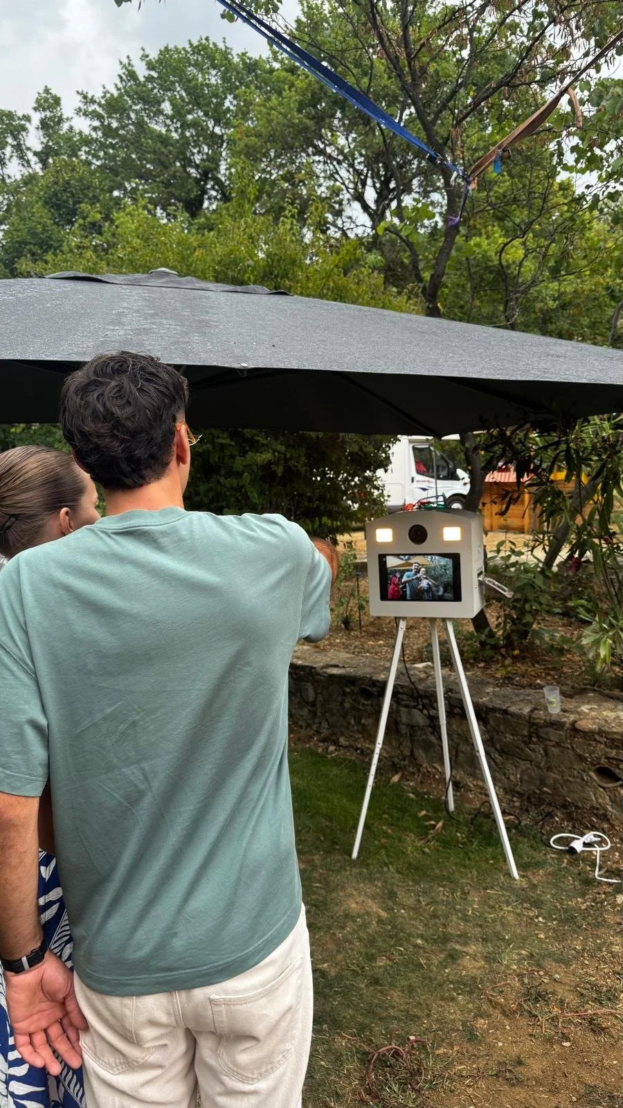 People standing under an umbrella, looking at a camera on a tripod that is capturing their reflection.