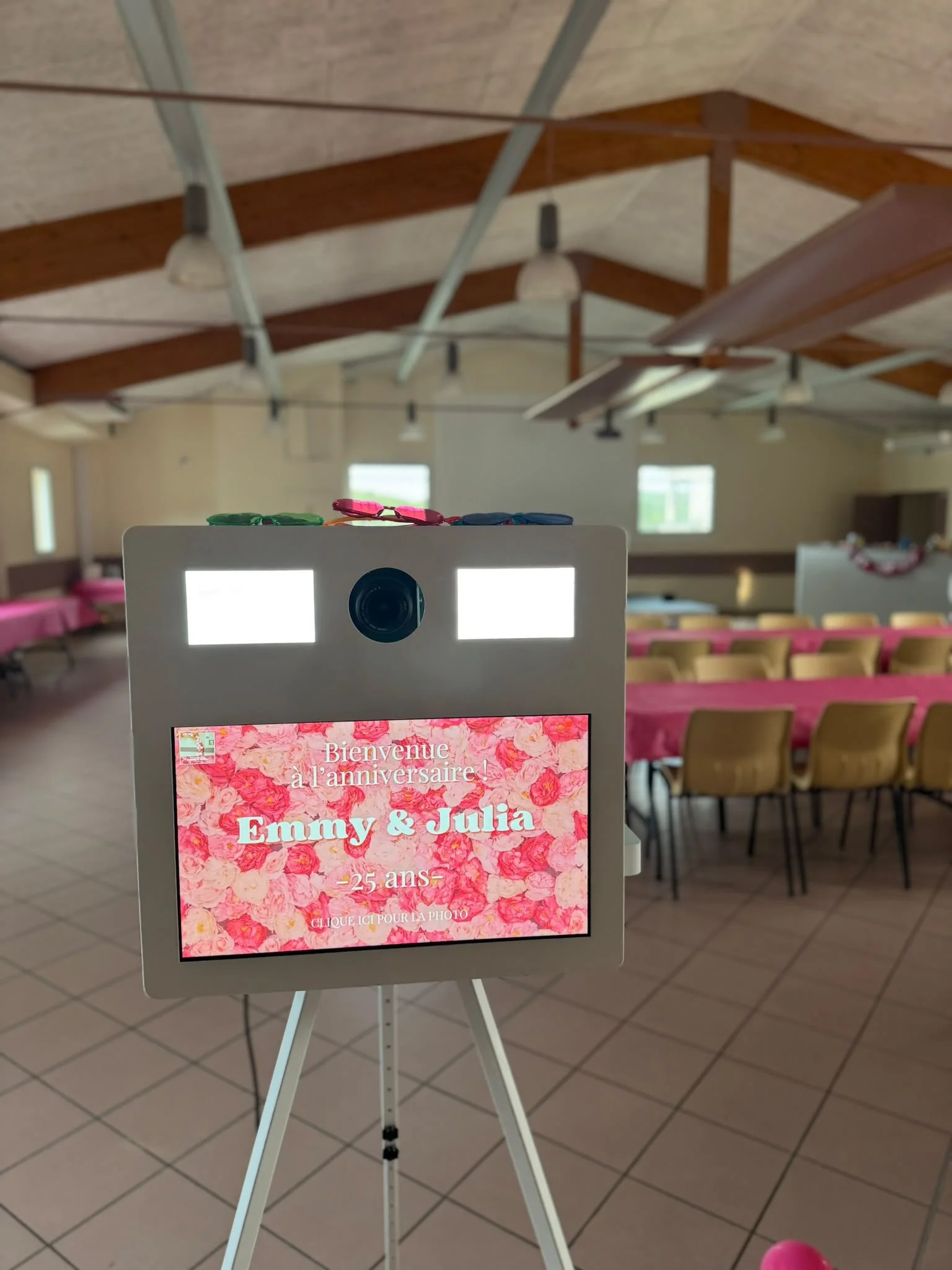 A sign on a stand in a large room with rows of chairs and pink tablecloths, welcoming Emily and Julia for their 25th birthday celebration with a floral background.