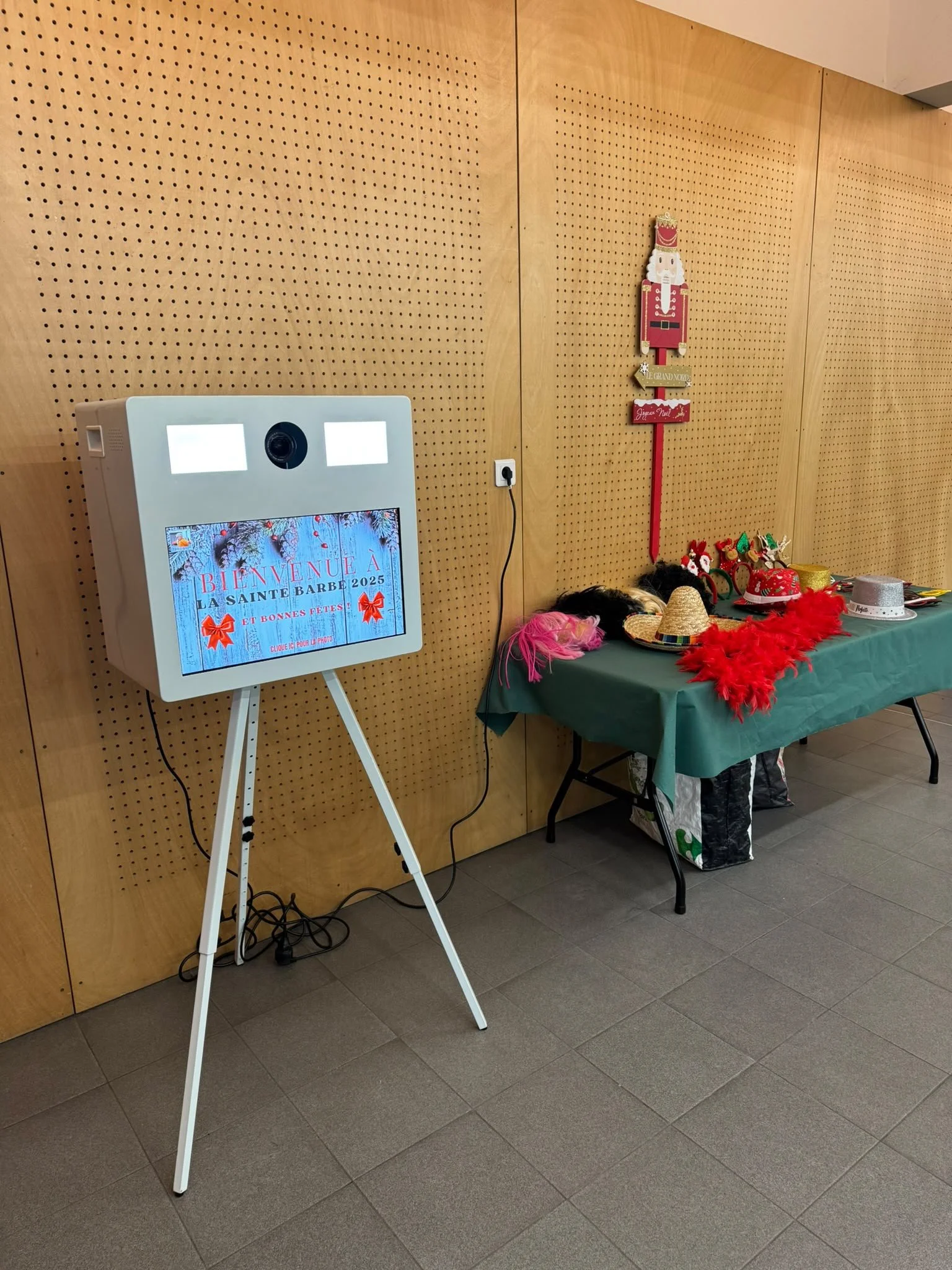 A photo of a wall with a table on the right side covered with Christmas items like hats, feather boas, and accessories. To the left, there is a white electronic photo booth or kiosk with a screen displaying a holiday greeting in French, a camera, and two blank rectangles, possibly lights or indicators. A power outlet with an extension cord is also visible.