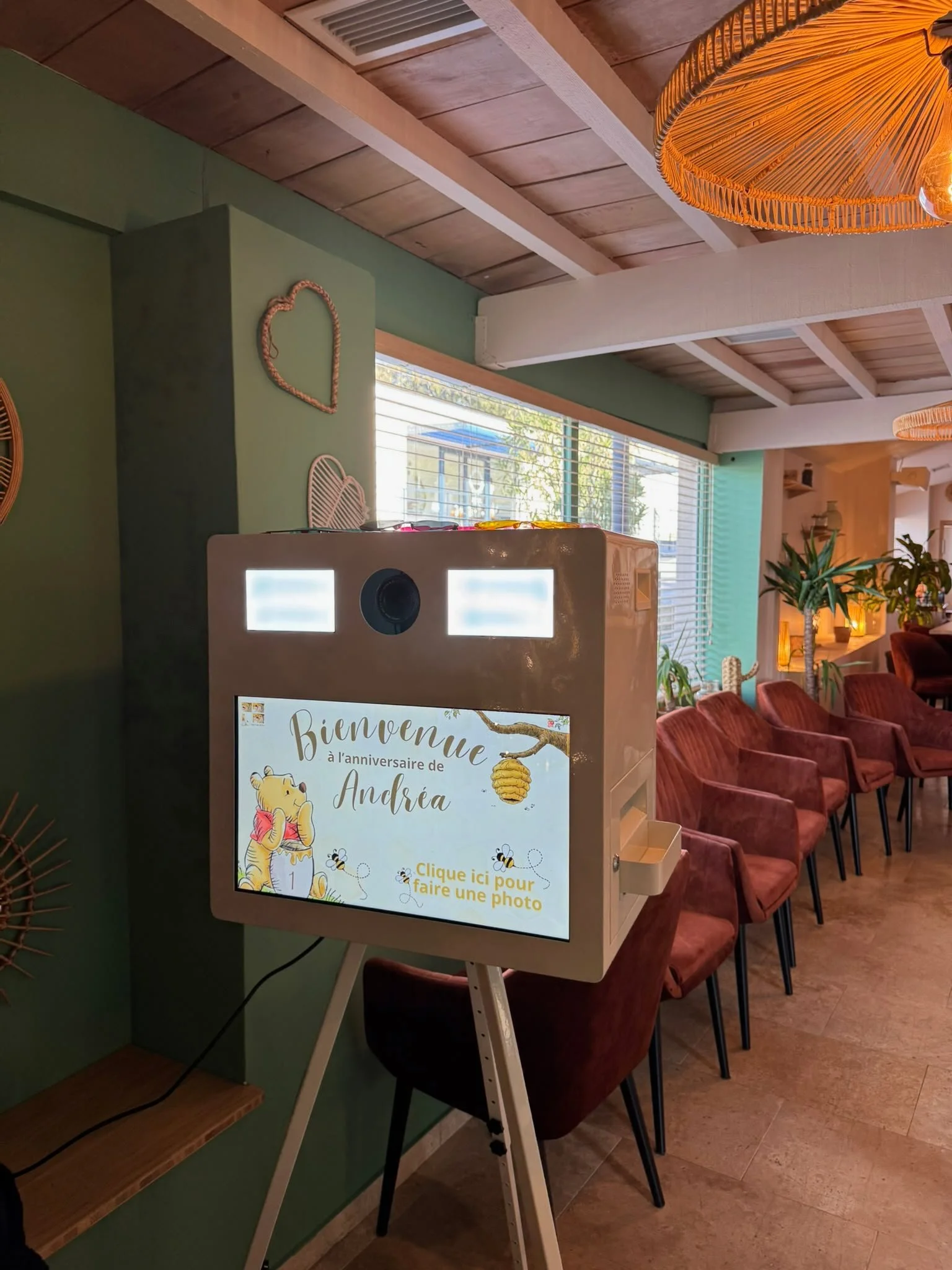 A photo booth with a greeting message for Andrea's birthday featuring Winnie the Pooh and bees, situated in a room with green walls, wooden ceiling, and natural light from large windows. Pink chairs are arranged in rows nearby and hanging light fixtures are also visible.