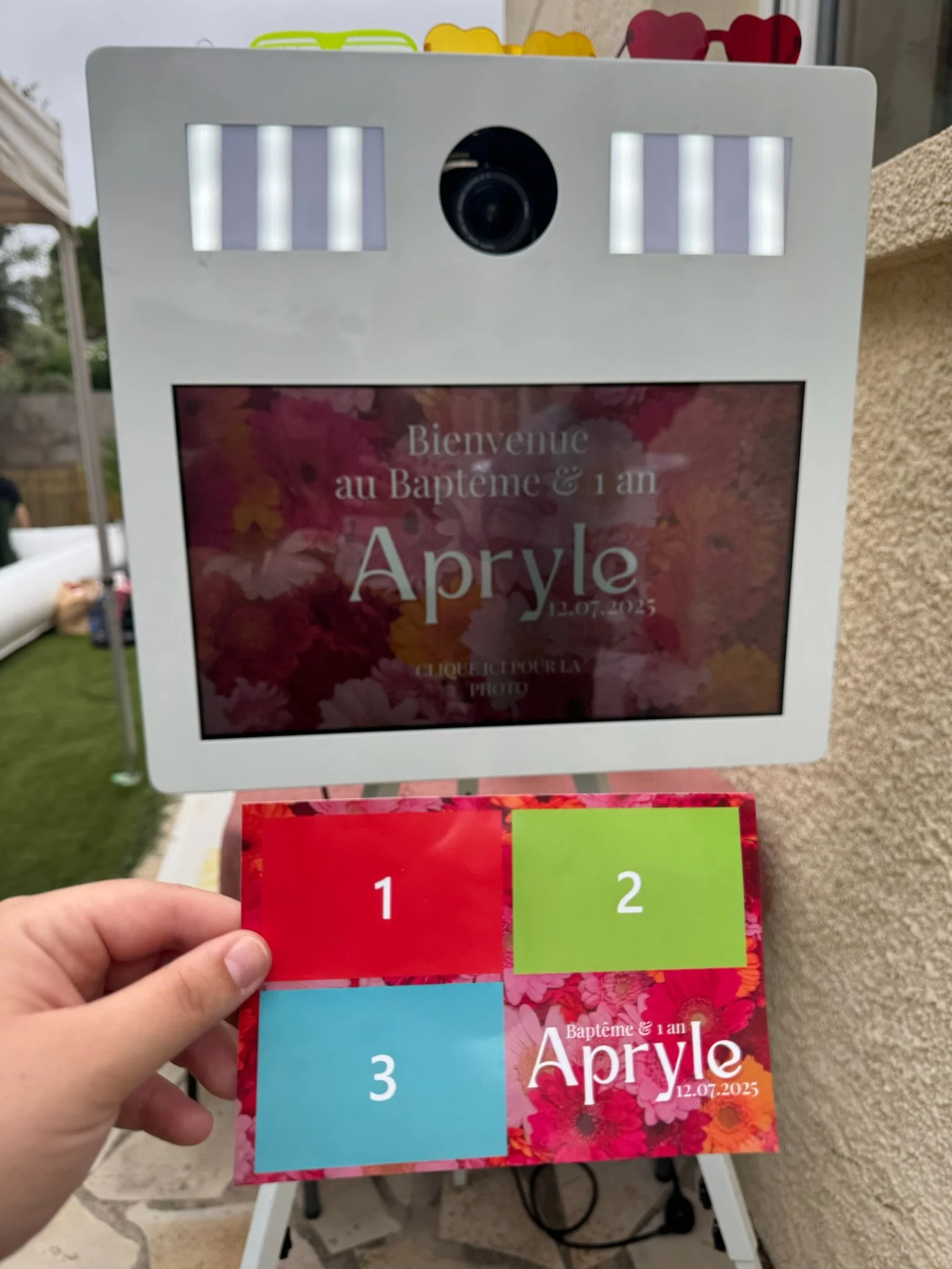 Une personne qui tient une impression photo devant la borne photo. 
L’écran de la cabine photo affiche le message de l'évènement. 