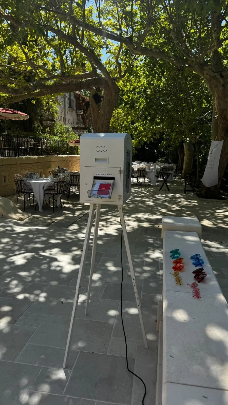 A white voting ballot box on a tripod stand outdoors under a large tree with green leaves. A small table nearby holds colorful clothespins. In the background, there are tables with white tablecloths and chairs at a patio restaurant or café.