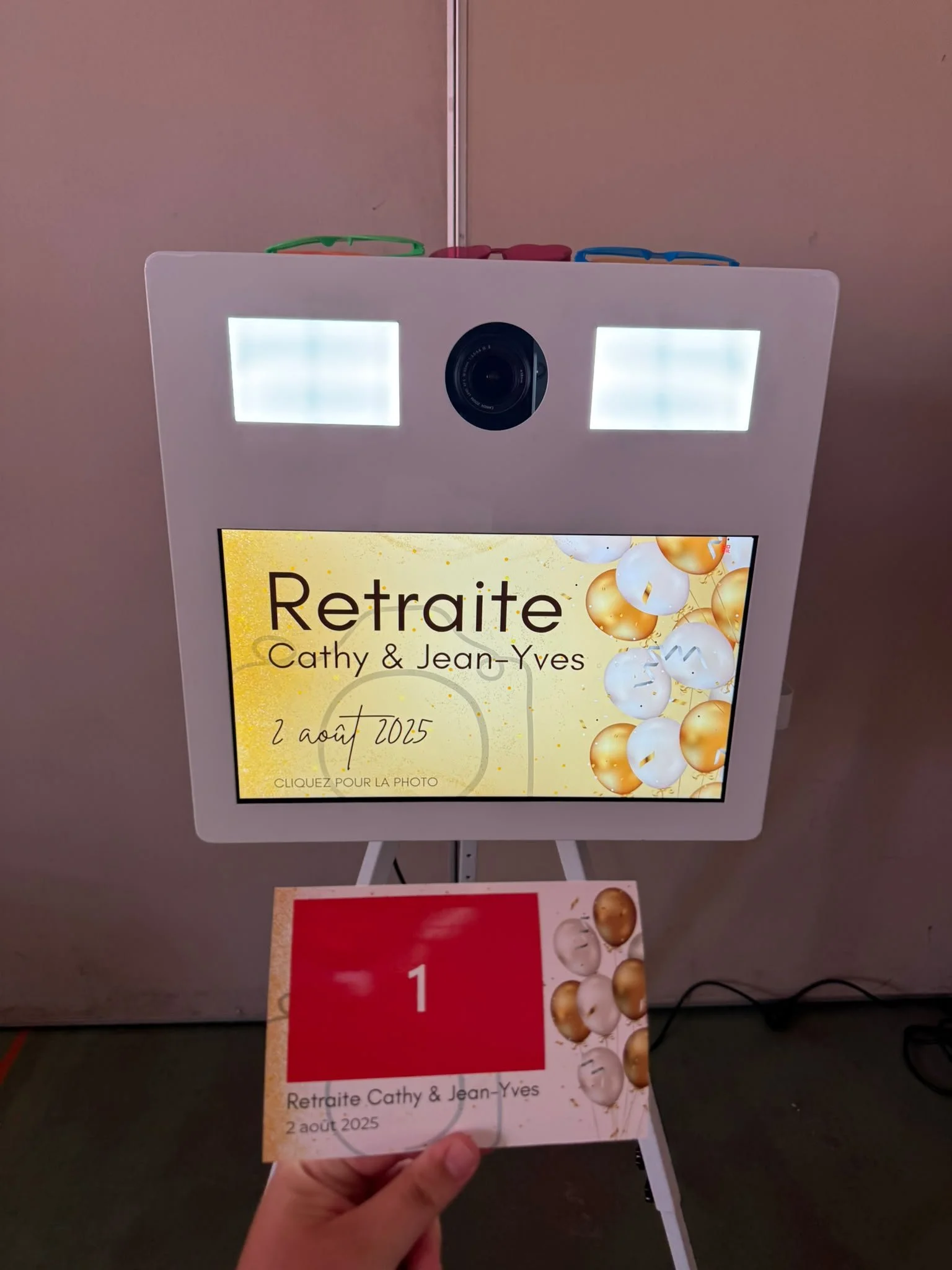 Digital photo booth screen displaying invite for retirement celebration for Cathy and Jean-Yves on August 2, 2025, with a decorative background of white and gold spheres.
