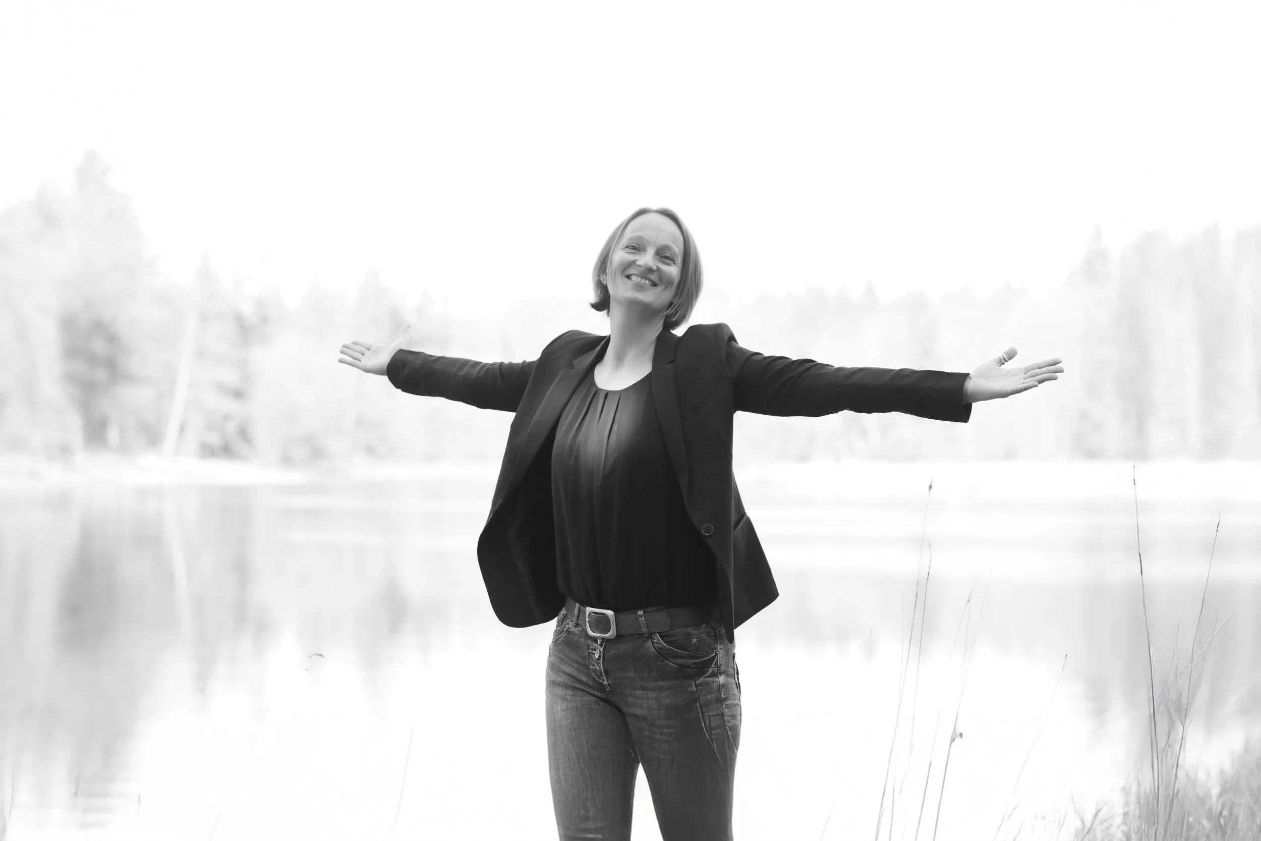 Lächelnde Frau (Simone Niederl) mit offenen Armen vor einem Fluss, im Freien, in Schwarz-Weiß.