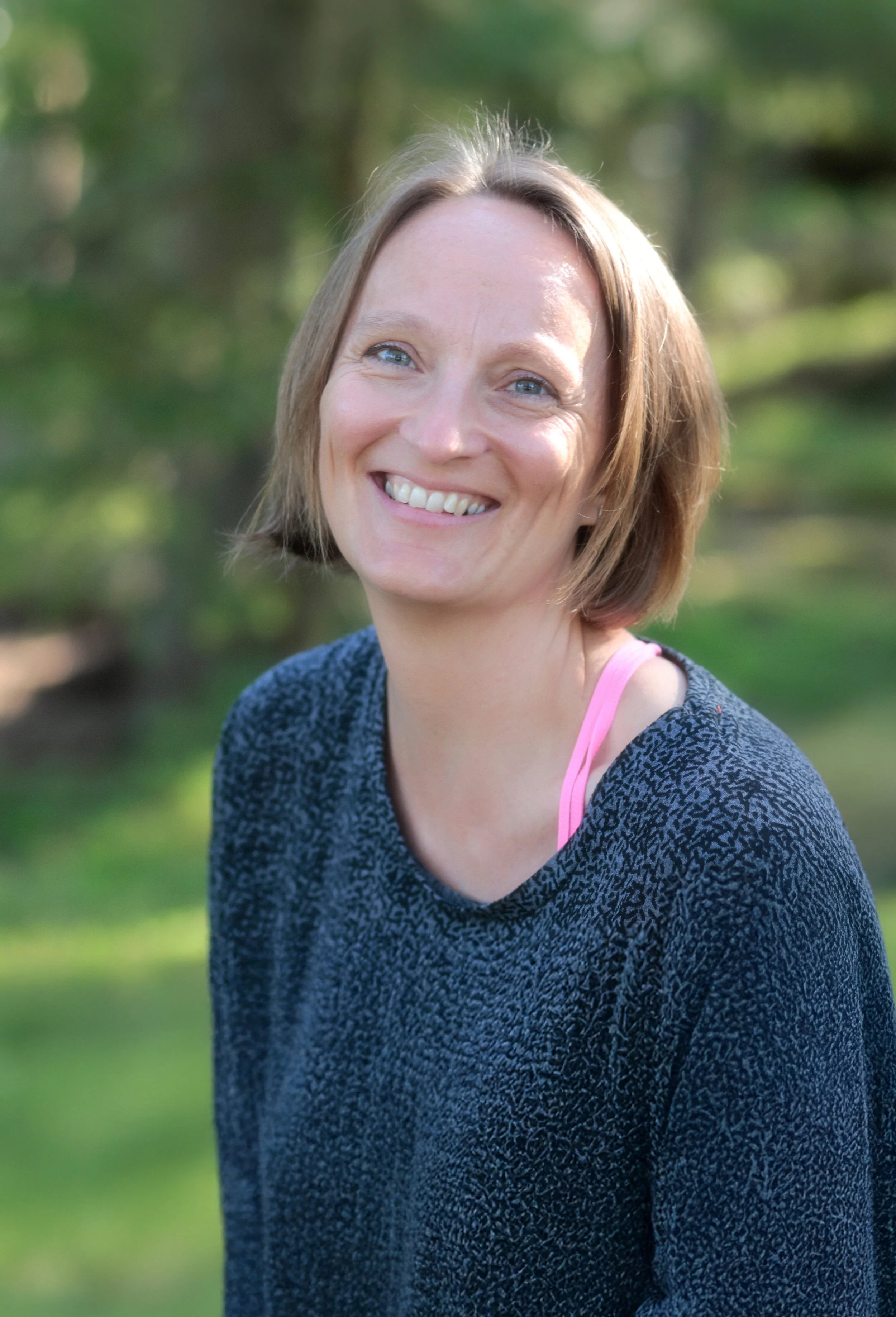 Lächelnde Frau (Simone Niederl) mit kurzen Haaren, trägt einen dunkelblauen Pullover und einen pinken Sport-BH, steht im Grünen.