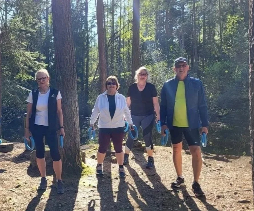 Vier ältere Menschen stehen in einem Wald, alle tragen sportliche Kleidung. Es sieht aus, als ob sie gerade Sport gemacht haben.