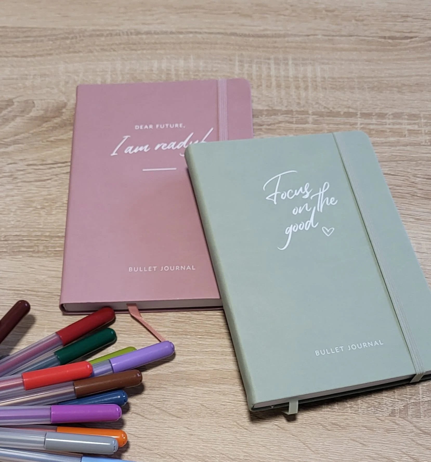 Zwei Bullet Journals, einer pink und einer hellgrün, liegen auf einem Holztisch, umgeben von bunten Filzstiften. Die pinke Titelseite trägt die Aufschrift 'Dear future, I am ready' und die grüne 'Focus on the good'.