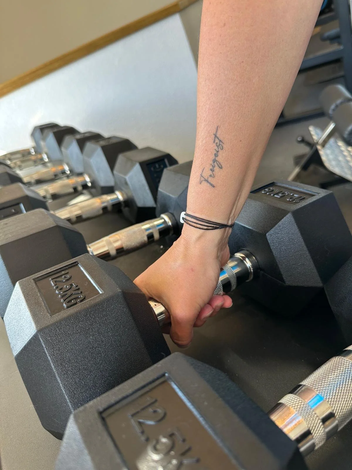 Hand greift nach einer Hantel im Fitnessstudio, mit Tattoo 'Freigeist' am Unterarm.