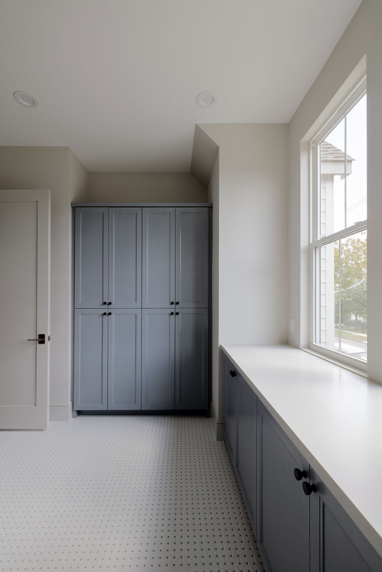 Laundry room with tall blue-gray storage cabinets, window-side folding counter, and basketweave mosaic tile