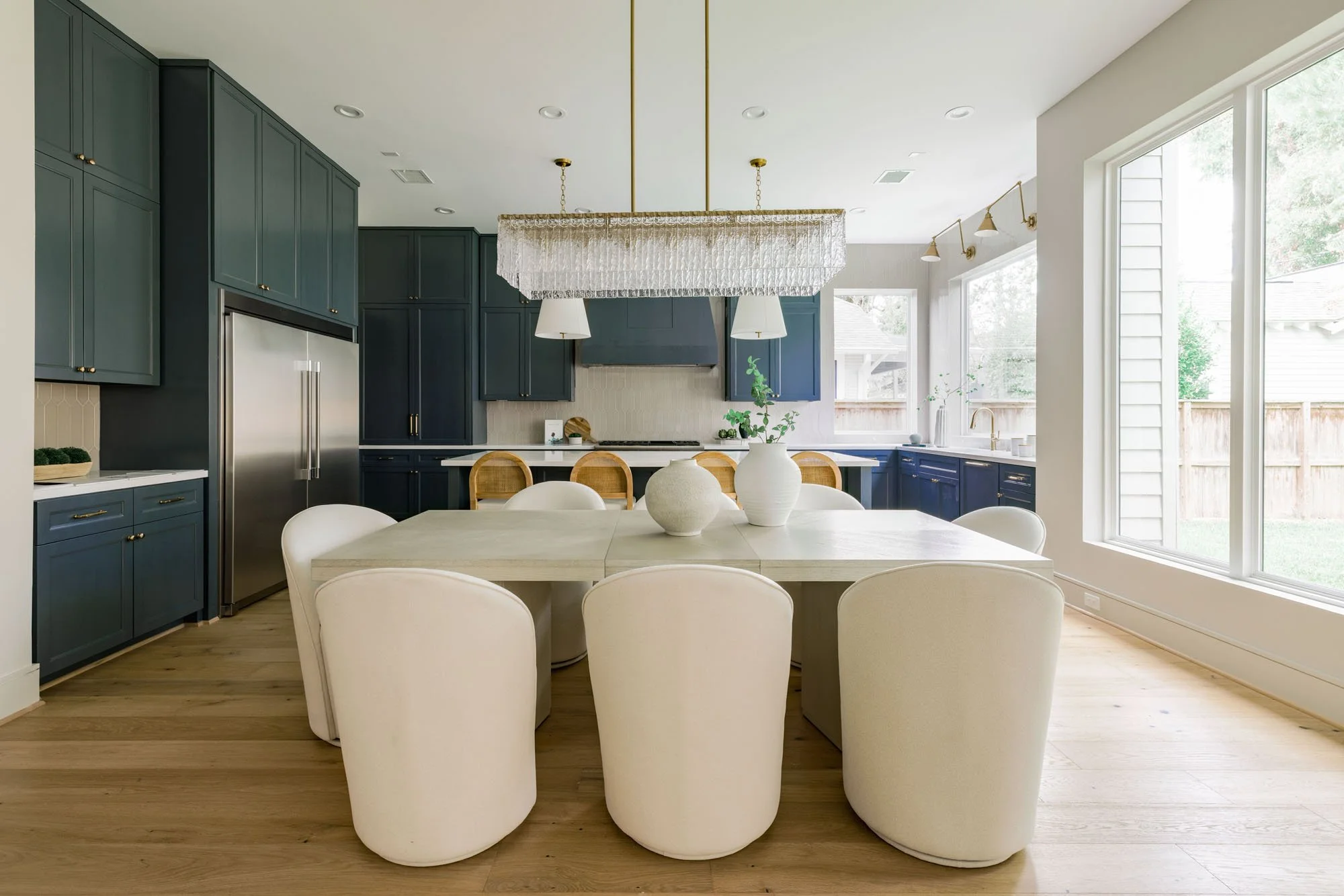 Dining area with ivory swivel chairs and crystal chandelier facing a navy blue kitchen with built-in fridge