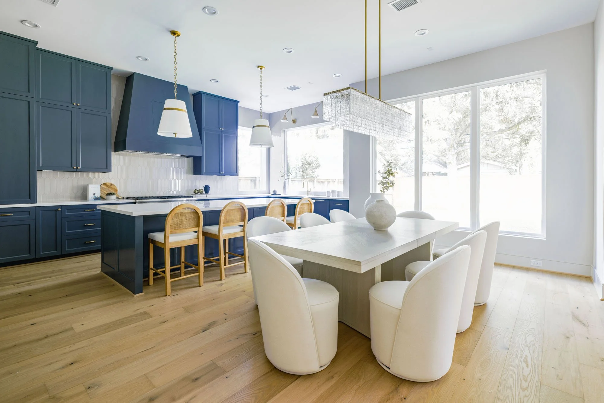 Open kitchen with navy cabinets, rattan counter stools, and dining table under a glass chandelier