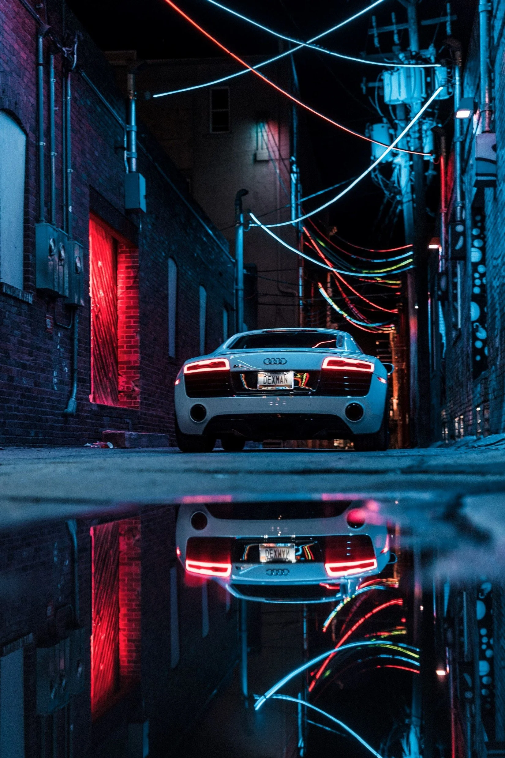 A silver Audi sports car parked in a dark alley with colorful neon lights and wires overhead, reflected in a puddle on the ground.