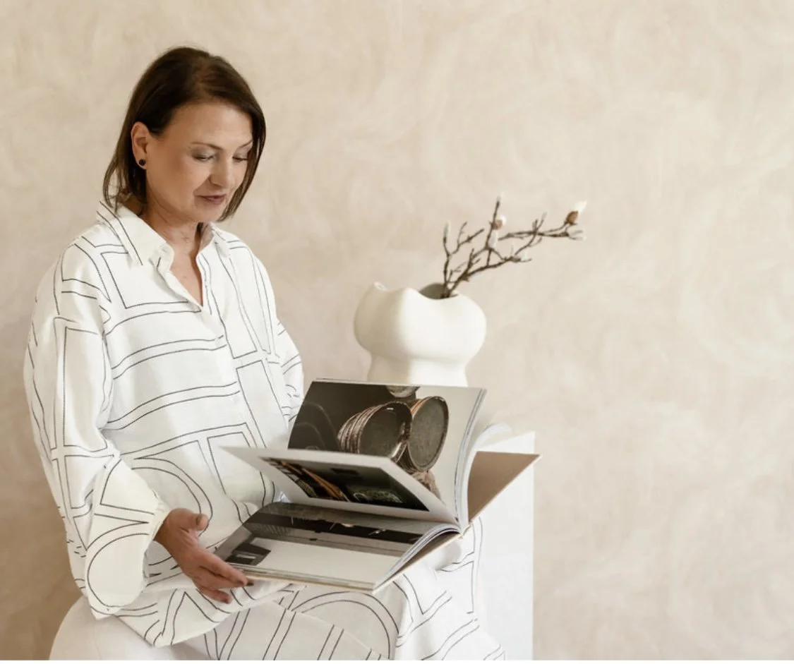 Frau liest ein Magazin in einem weißen Raum mit einer weißen Vase mit Ästen im Hintergrund.