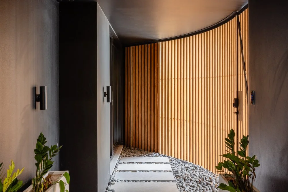 Modern outdoor corridor with a curved wooden wall, concrete flooring, and pebble stones, decorated with potted plants.