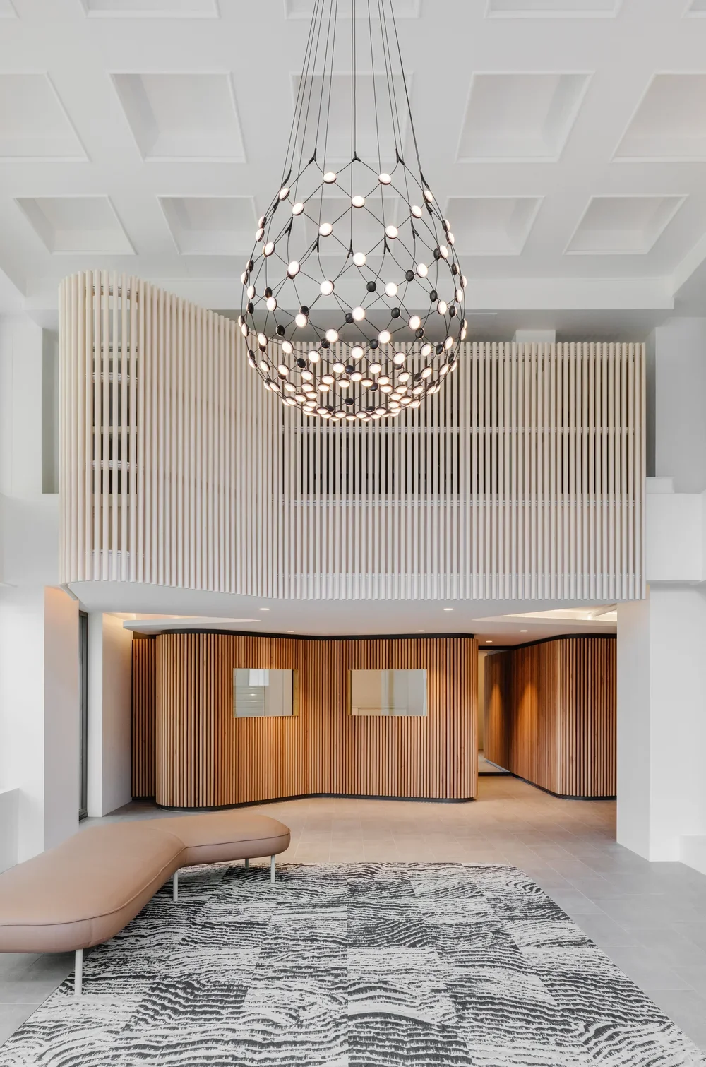 Modern lobby featuring a large, spherical chandelier with multiple lights, a beige curved sofa, a patterned rug, and wooden slat walls.