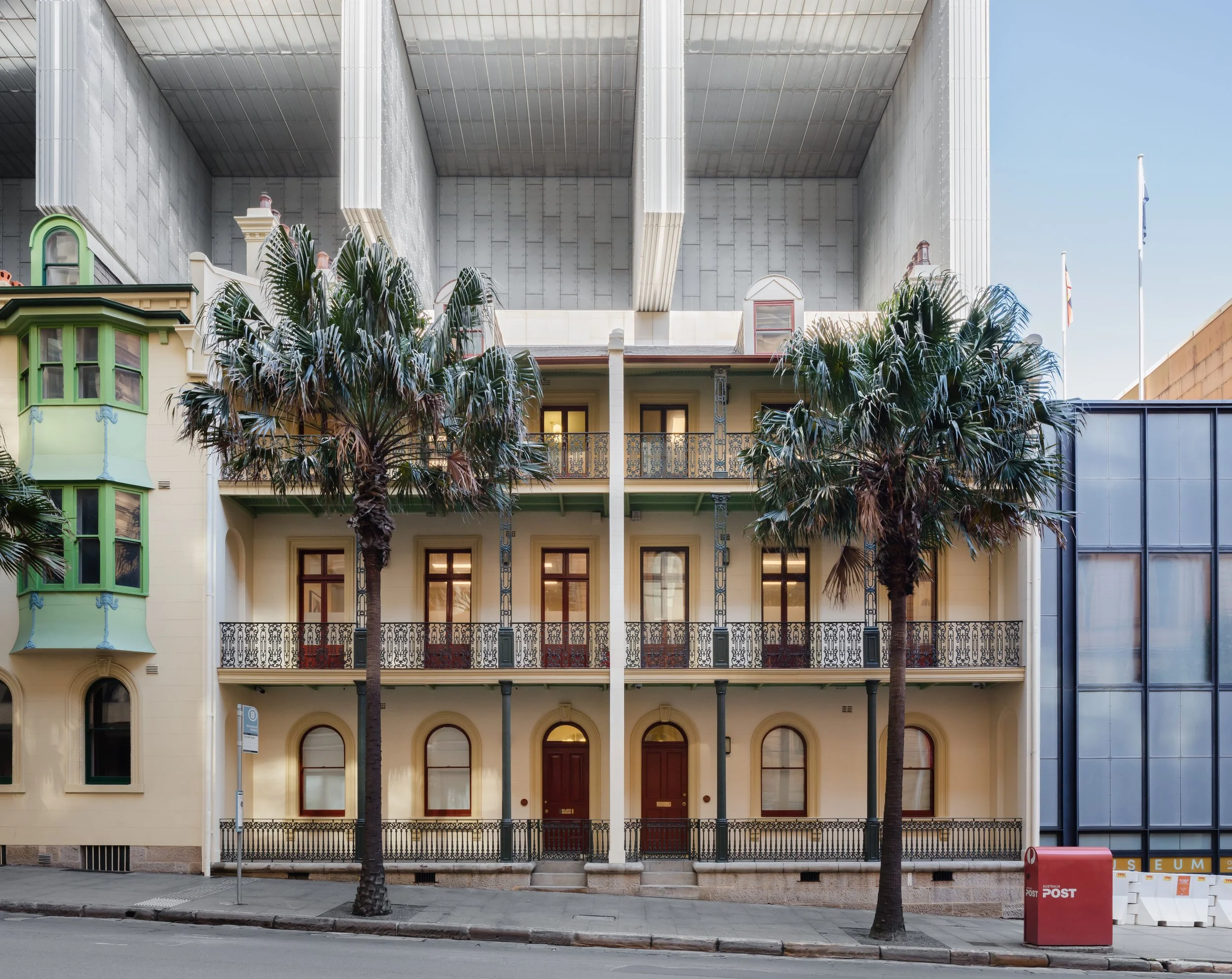 A historical brick building with two stories, featuring a decorative wrought-iron balcony and arched windows and doors, flanked by two palm trees on a city street with a mailbox and parking sign.