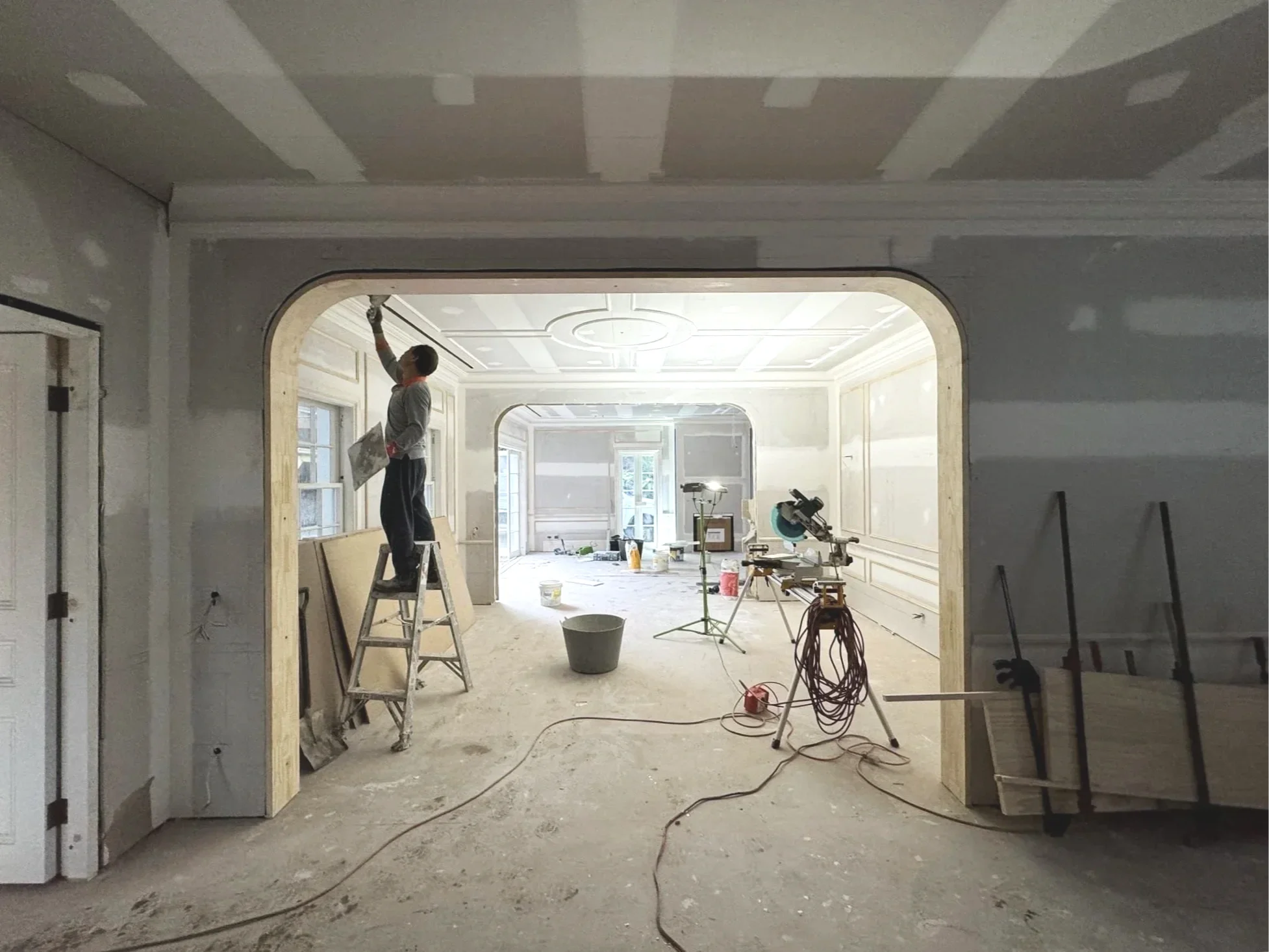 A person standing on a ladder painting a ceiling in a room under construction, with construction tools and materials scattered around.