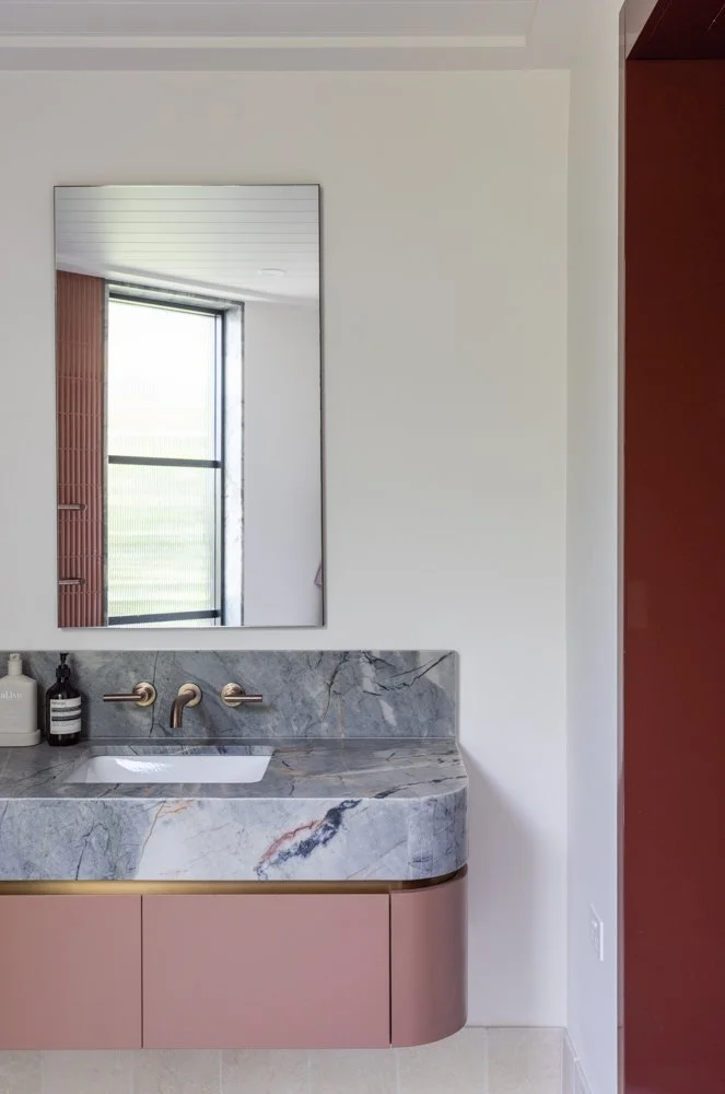 Modern bathroom vanity with a marble countertop, pink cabinet, and a mirror above. Reflection shows a window with a view outside.