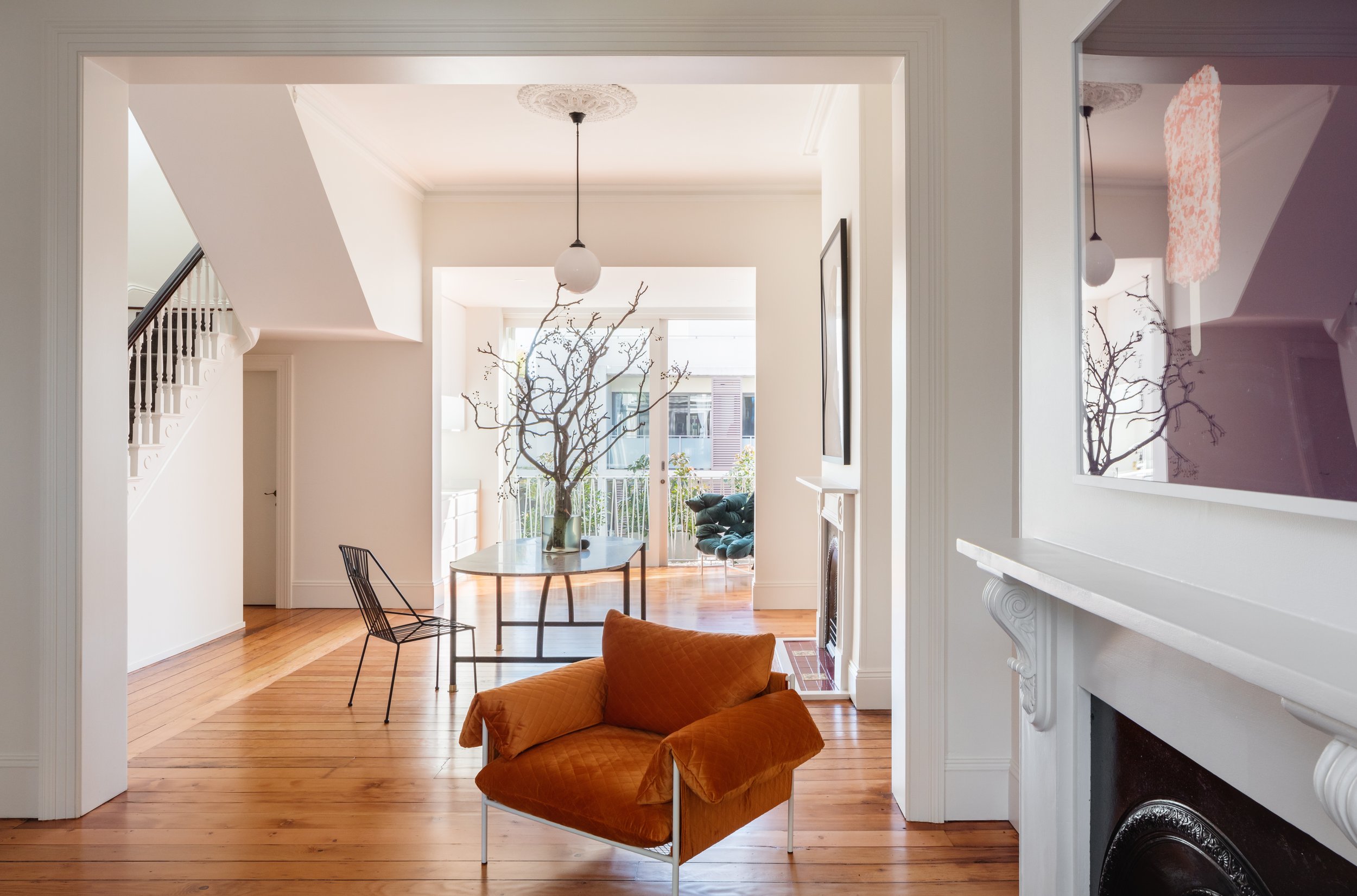 Bright living room with hardwood floors, a white fireplace, an orange armchair, and a modern glass table with a branch arrangement, leading to a balcony with sliding glass doors.
