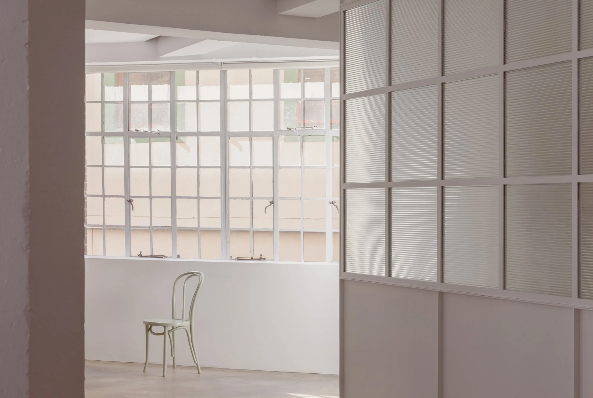 A white chair near a large window with multiple small panes and locks, inside a minimalist room with white walls and a frosted glass partition.