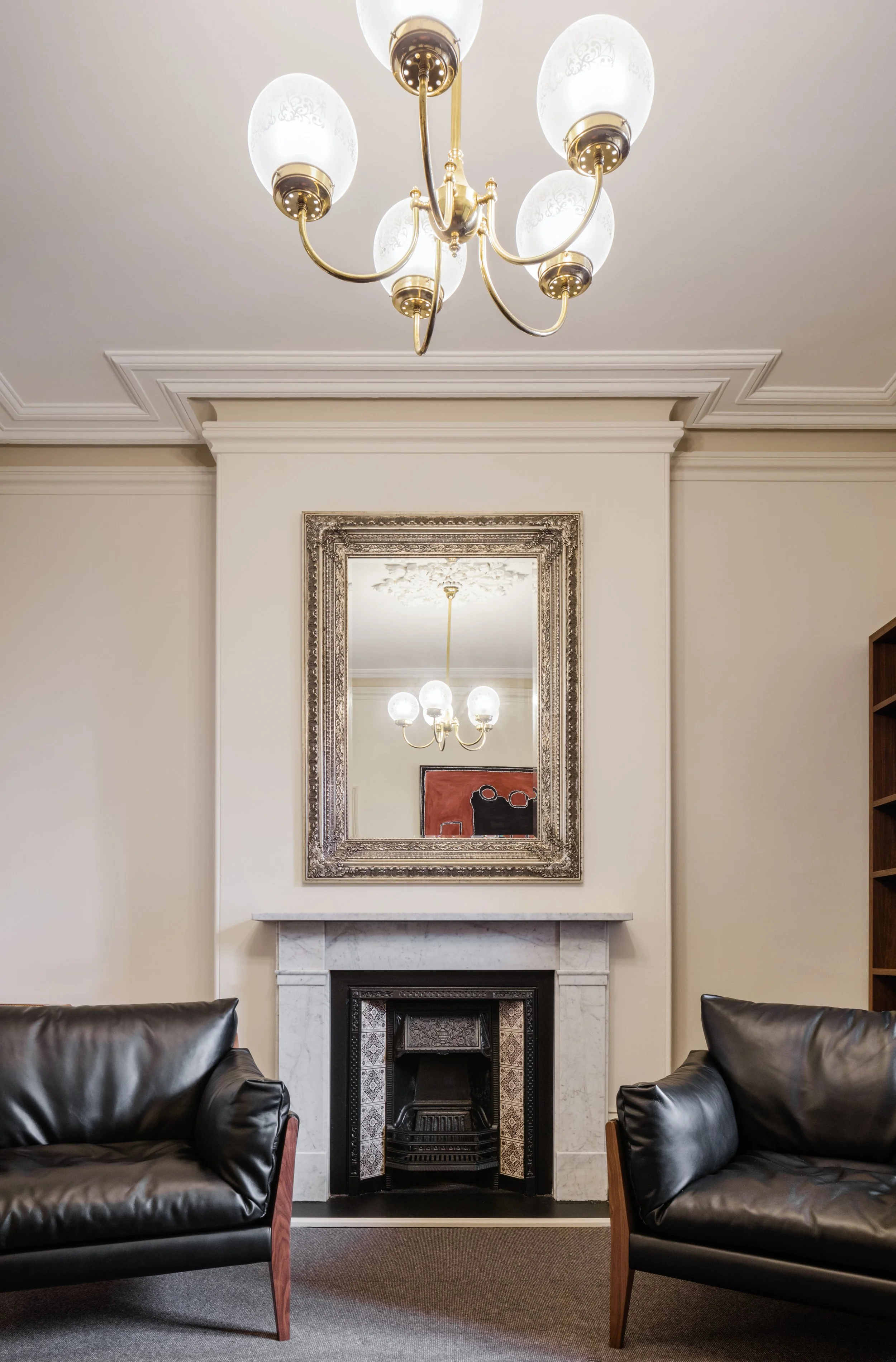 Living room with beige walls, a white marble fireplace, a large ornate silver-framed mirror above the fireplace, a chandelier hanging from the ceiling, and two black leather armchairs facing each other.
