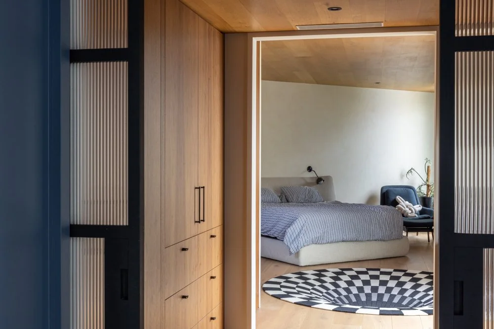 View of a modern bedroom through an open doorway, featuring a bed with gray bedding, a black armchair with a teddy bear, and a patterned rug on light wooden floors.