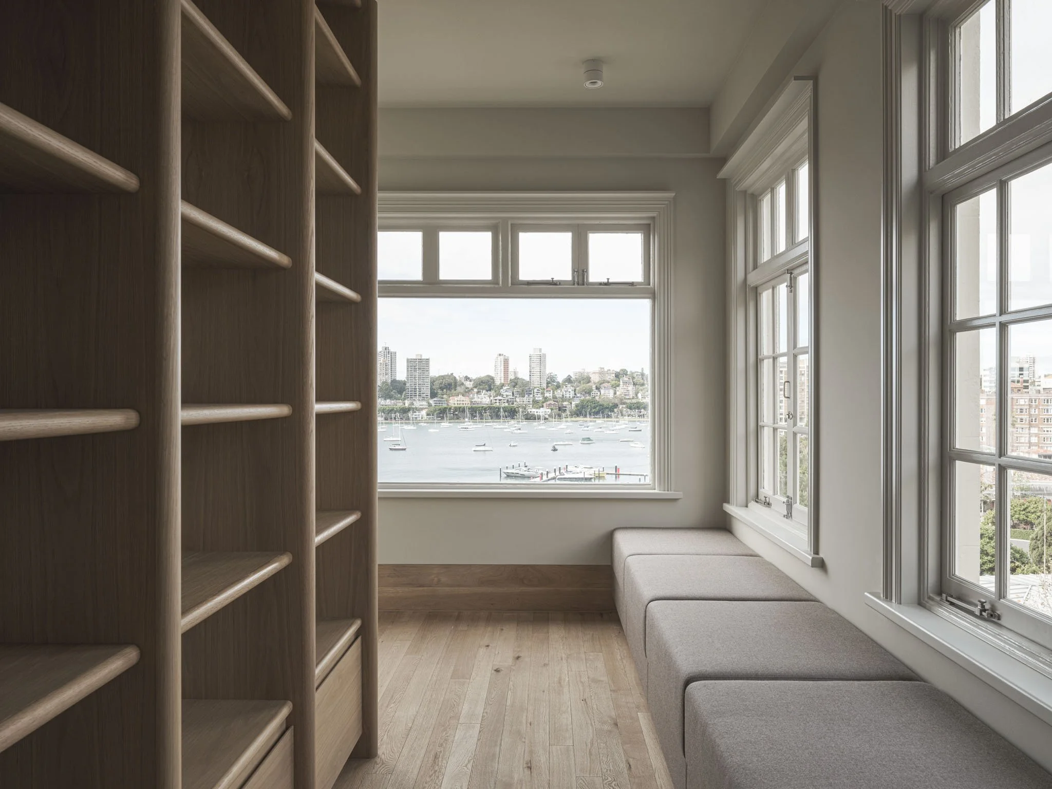 Room with large windows overlooking a city and water view, with a built-in bench and wooden shelving on one side