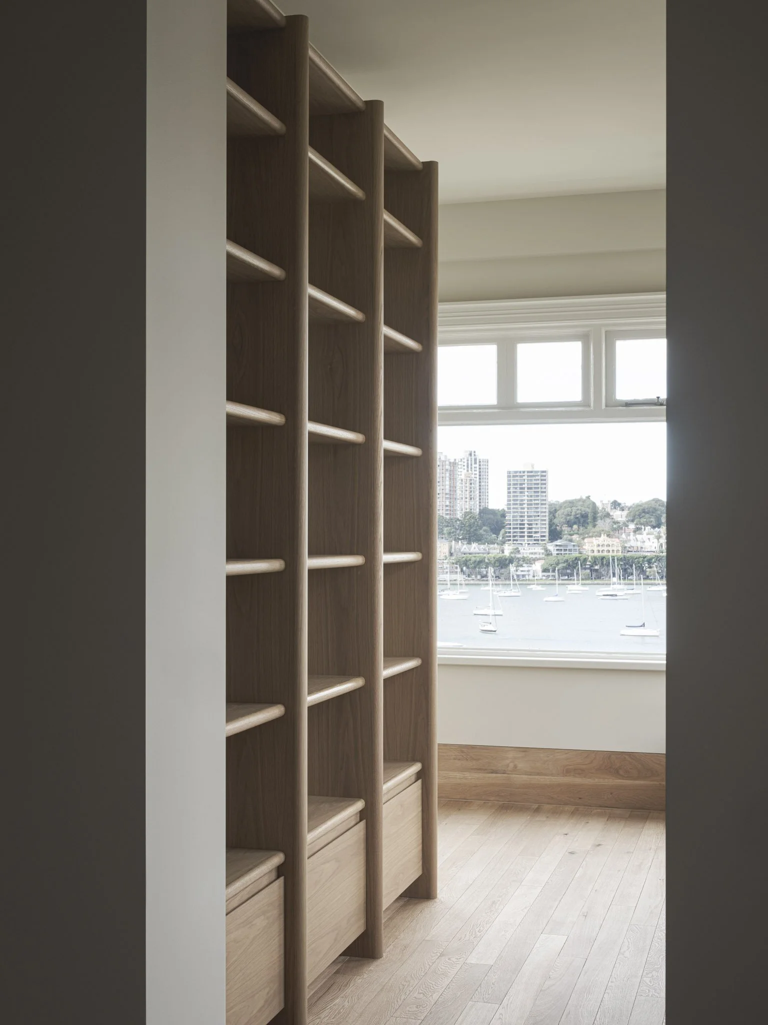 Tall wooden bookshelf with open shelves situated in a room with large window overlooking a harbor with boats and cityscape.