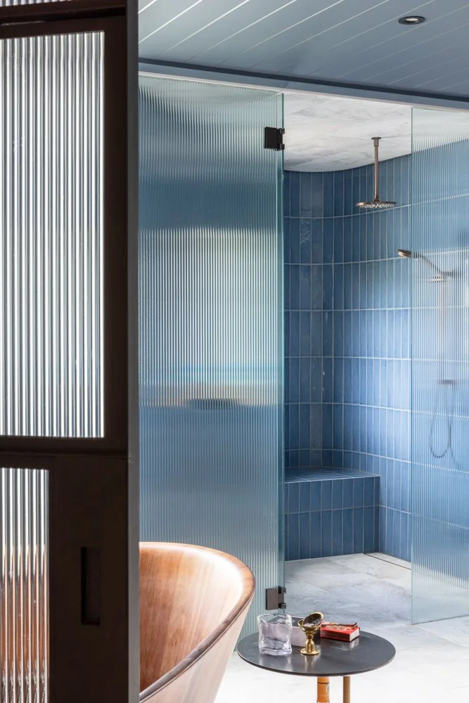 Modern bathroom with blue tiled shower area, glass partition, wooden bathtub, and small round black side table with a water jug, gold cup, and a red book.