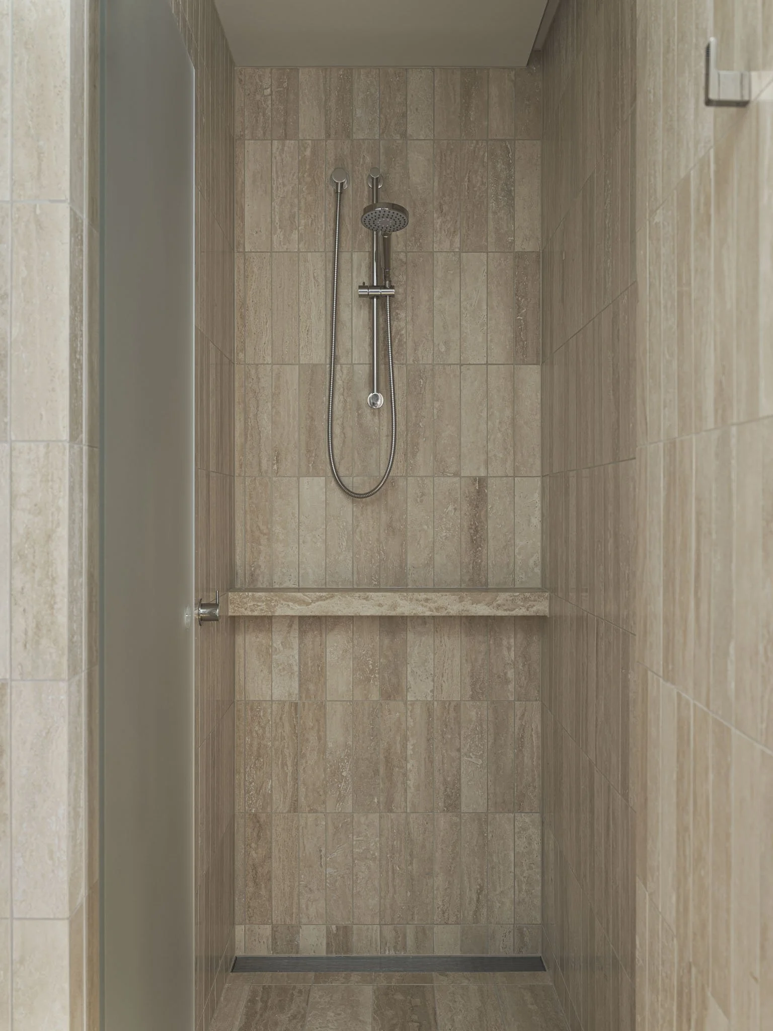 A walk-in shower with beige tiled walls, a silver handheld showerhead, a fixed showerhead, and a long horizontal bench.