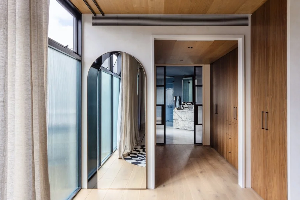Interior view of a modern room with large frosted windows, a mirror, wooden cabinets, a sliding glass door, and a glimpse of a kitchen with a marble island in the background.