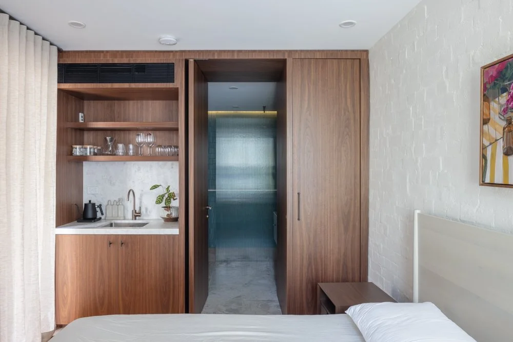 Hotel room with a bed, nightstand, small kitchenette with open shelves, glassware, and a plant, and a sliding wooden door leading to a bathroom with a frosted glass door.