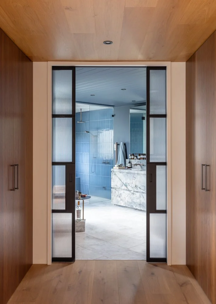 View through sliding glass doors into a modern bathroom with blue tiles, a white marble vanity, and a walk-in shower with a rain shower head.
