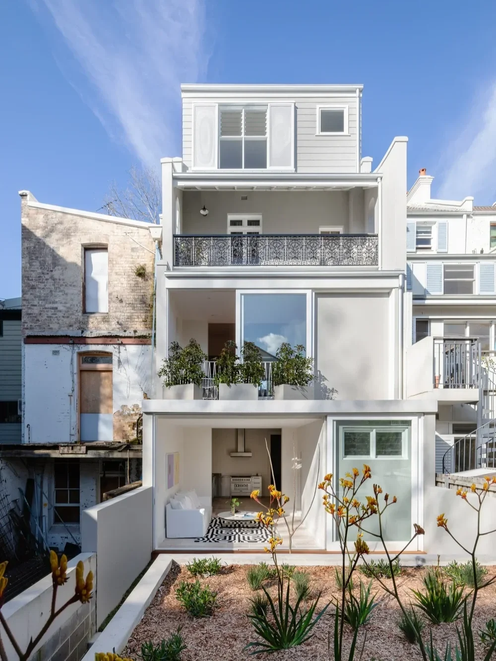 A modern white multi-story house with balconies, large windows, and a small front garden with plants and bushes, under a clear blue sky.