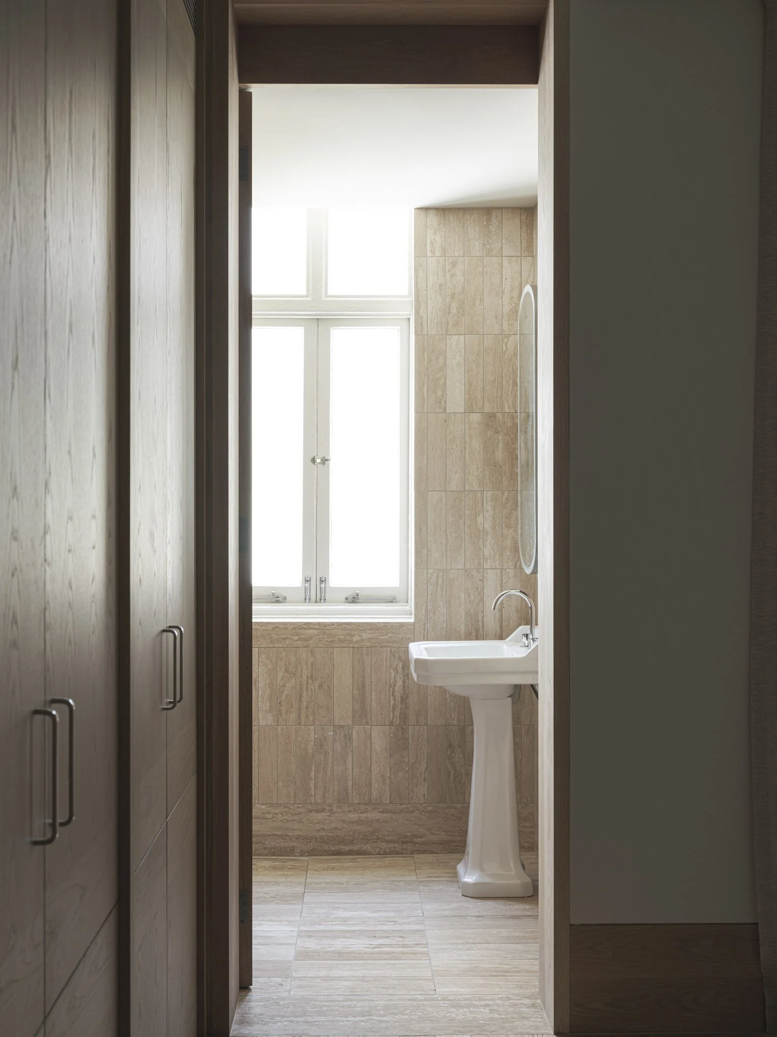 View into a small bathroom with a pedestal sink, a mirror, a window, and beige tiled walls and floor.